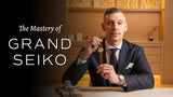 Man in a suit holds a Grand Seiko watch with a brown leather strap in a showroom setting