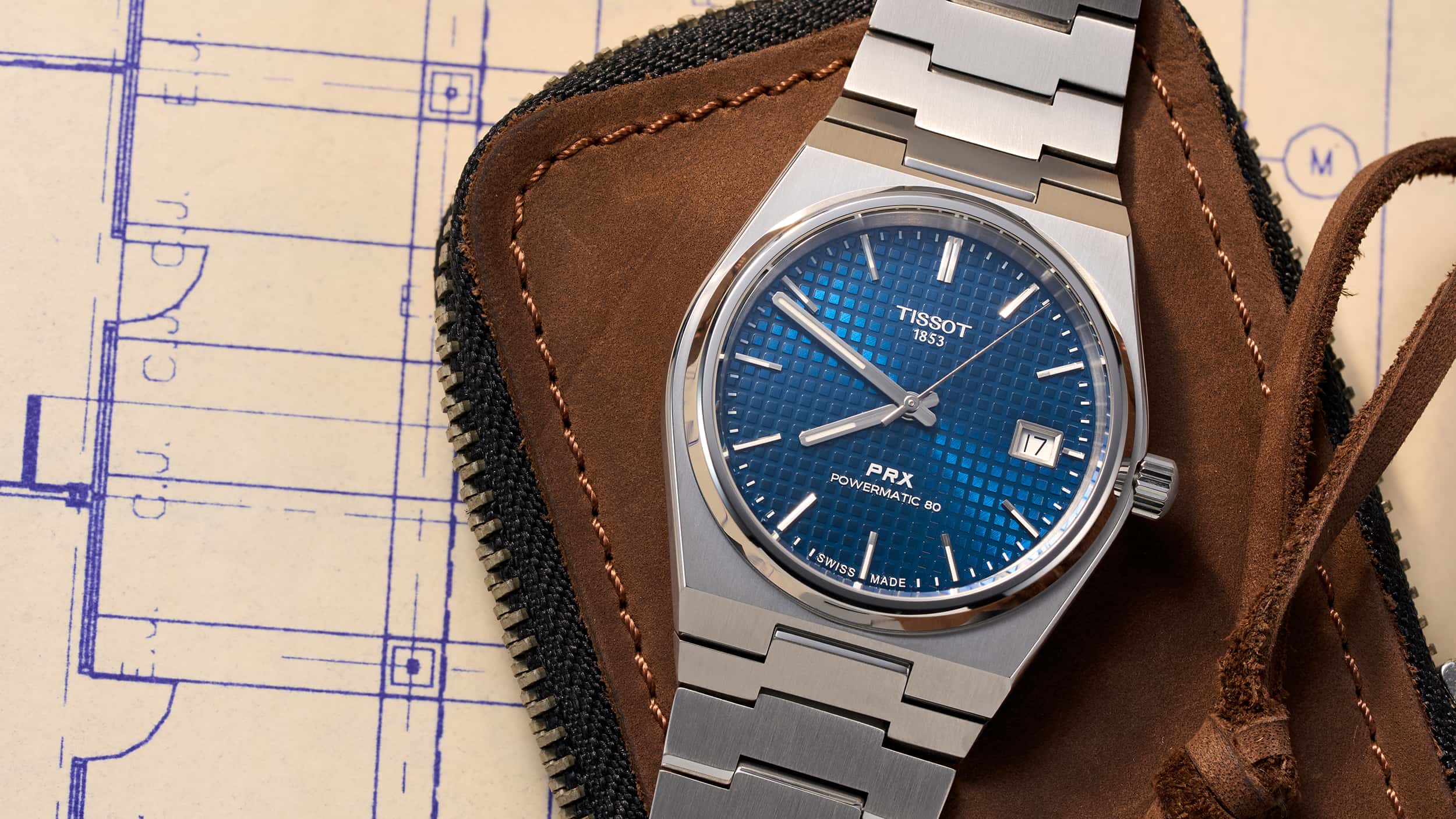 Close-up of a blue Tissot PRX Powermatic 80 wristwatch on a brown leather case with blueprint background.