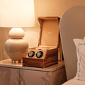 Open wooden watch winder on a nightstand with two watches inside under warm bedside light