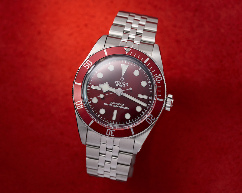 A TUDOR Swiss automatic watch with a silver stainless steel bracelet and case, featuring a dark red dial and a red and black rotating bezel, displayed on a textured red background.