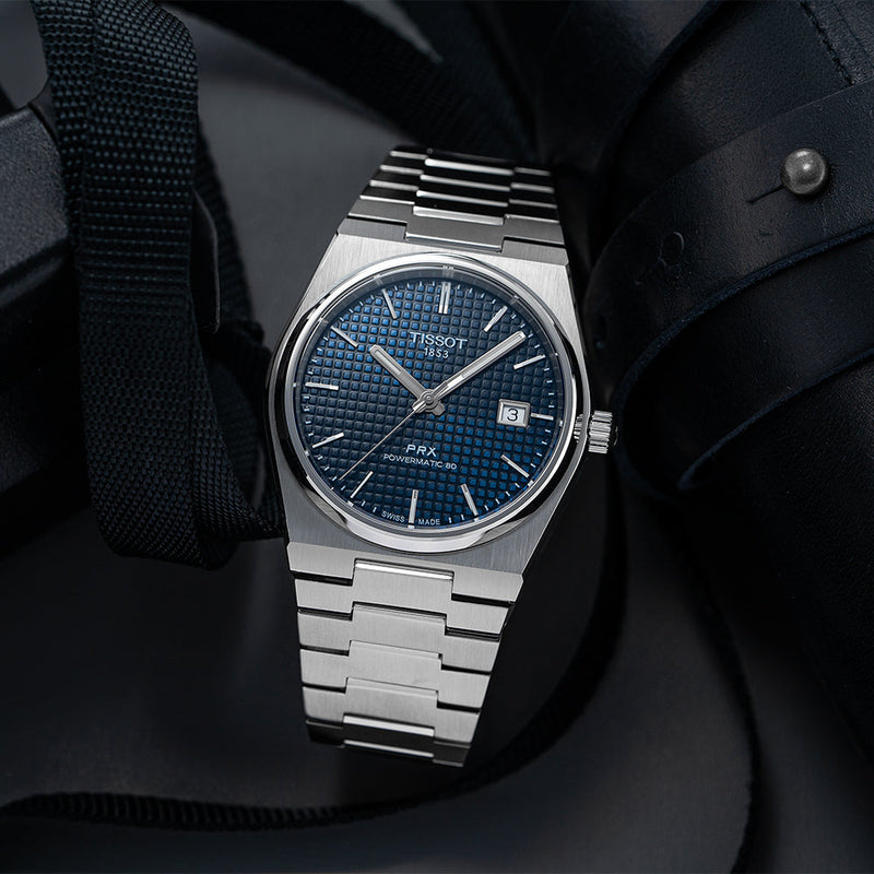 Tissot PRX Automatic Powermatic 80 blue dial watch on bracelet worn on the wrist against a dark backdrop